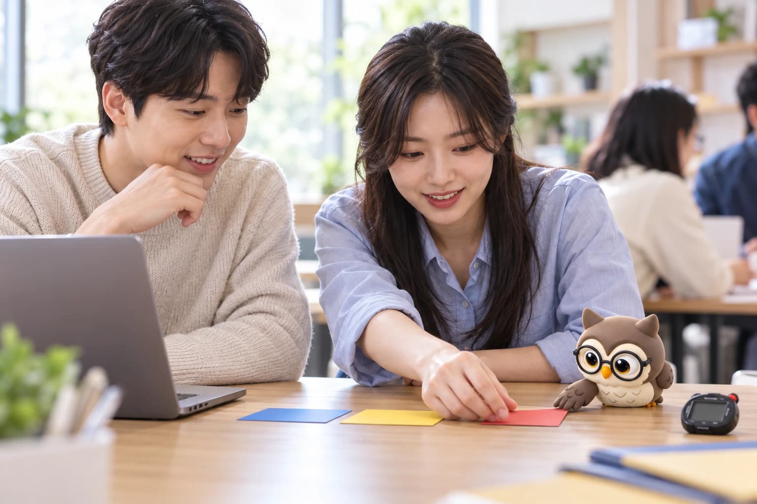 밝은 프로그래밍 학습실에서 두 사람이 색상 작업 카드를 배열하고 작은 갈색 안경 올리가 몸을 앞으로 기울여 날개로 카드를 미는 모습